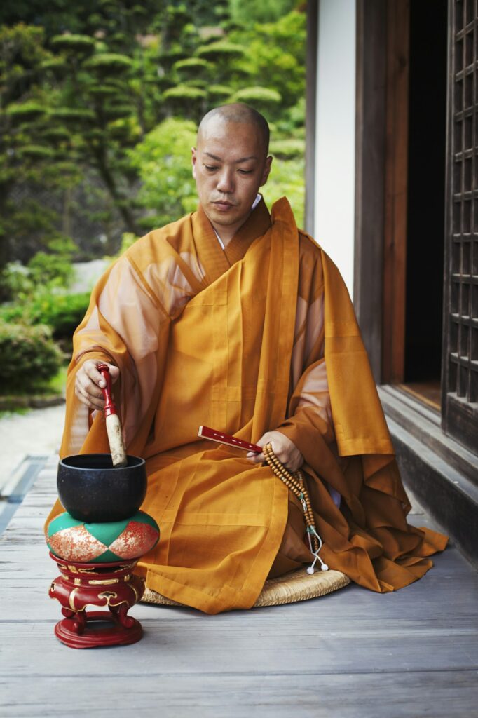 Buddhist monk with shaved head wearing golden robe sitting on floor outdoors, using singing bowl.